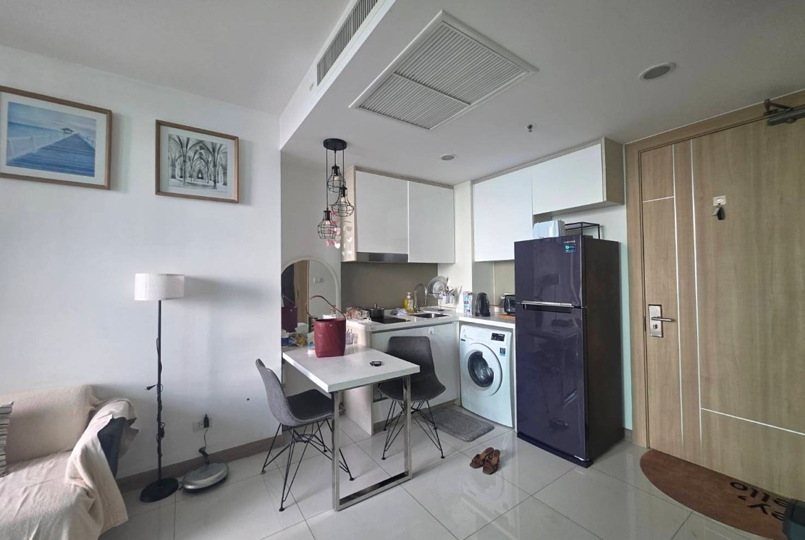 Cozy studio kitchen with white cabinets, a purple refrigerator, washing machine, dining table, and a sofa to the left; hanging pendant lights above the table.