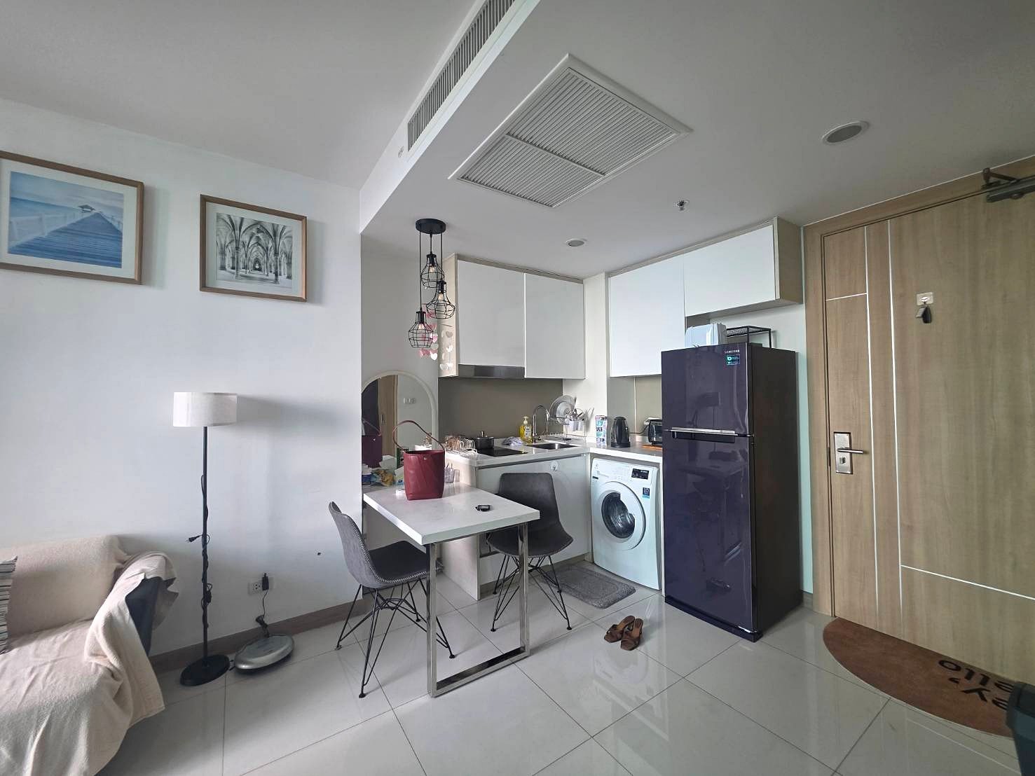 Riviera_Wongamat_B1209_2 Cozy studio kitchen with white cabinets, a purple refrigerator, washing machine, dining table, and a sofa to the left; hanging pendant lights above the table.