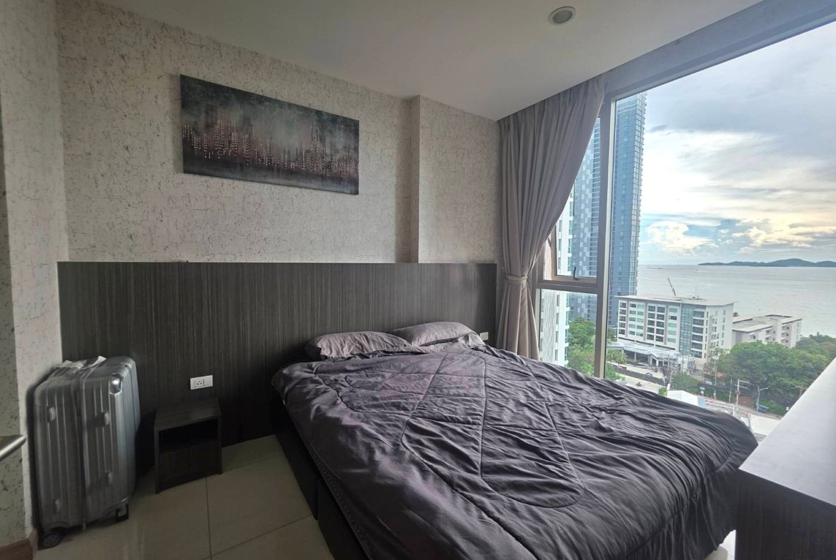 Queen bed with dark headboard and gray quilt in a modern room, large window showing a city and water view, suitcase beside the bed.