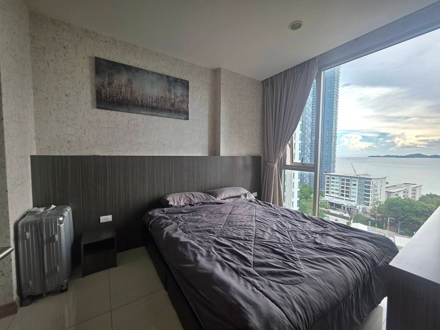 Riviera_Wongamat_B1209_3 Queen bed with dark headboard and gray quilt in a modern room, large window showing a city and water view, suitcase beside the bed.