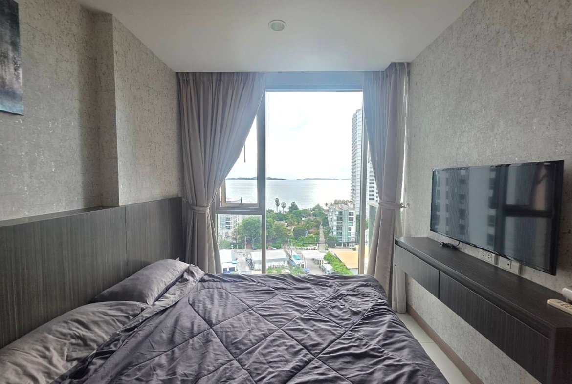 Bedroom with a grey quilted bedspread and a large window offering an ocean and city view far beyond the room.