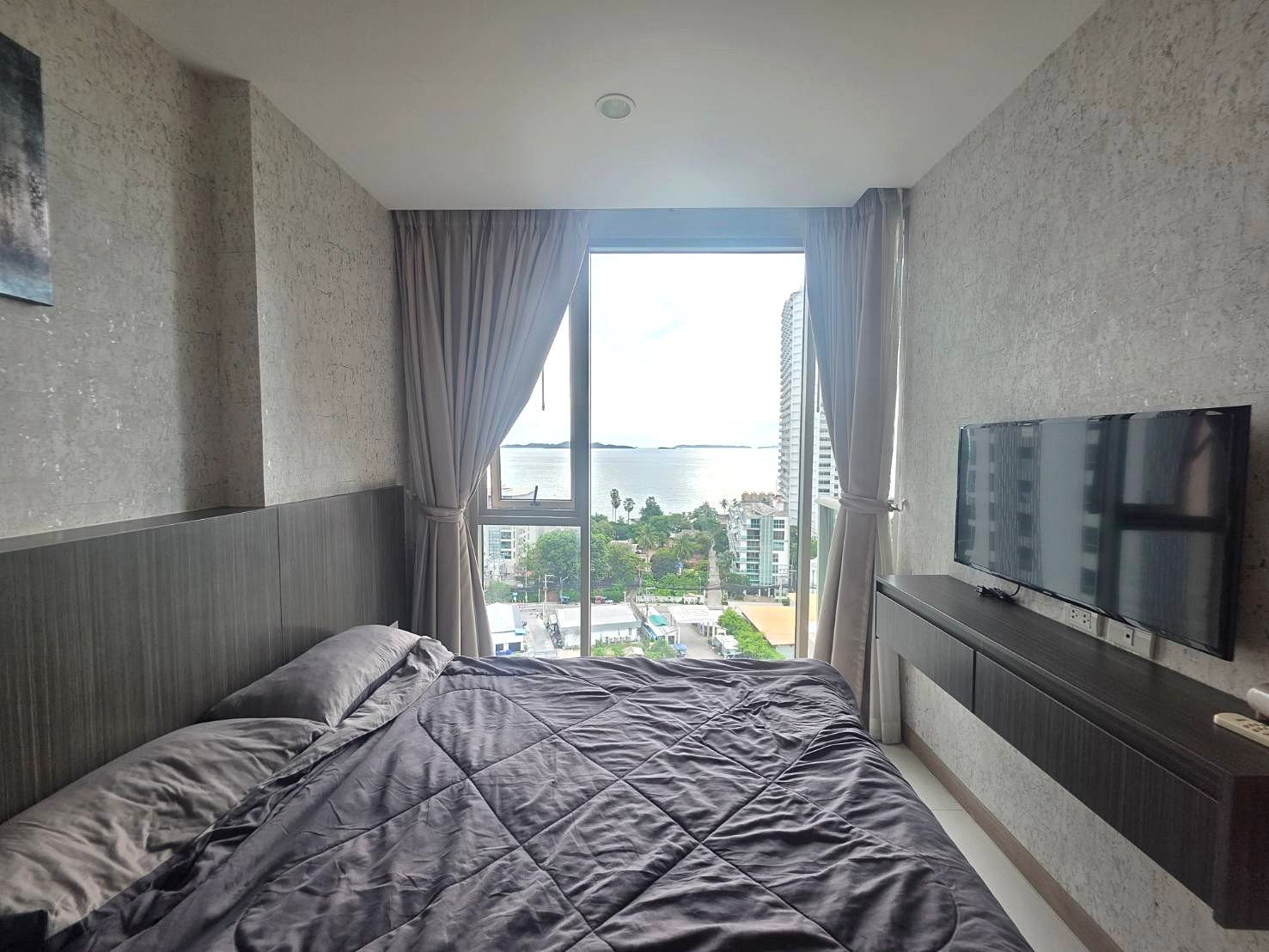Riviera_Wongamat_B1209_4 Bedroom with a grey quilted bedspread and a large window offering an ocean and city view far beyond the room.