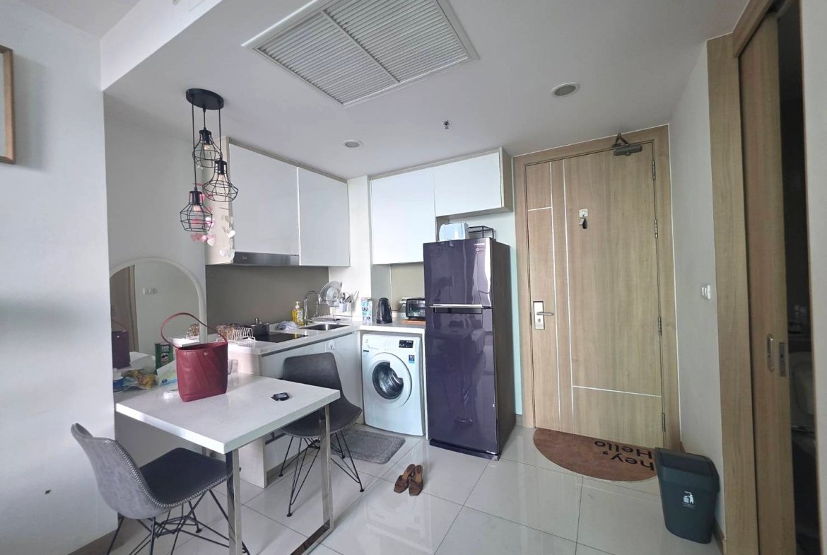 Compact open-plan kitchen with white cabinets, a purple fridge, washing machine, and a small dining table with two chairs, in a bright space with hanging pendant lights.