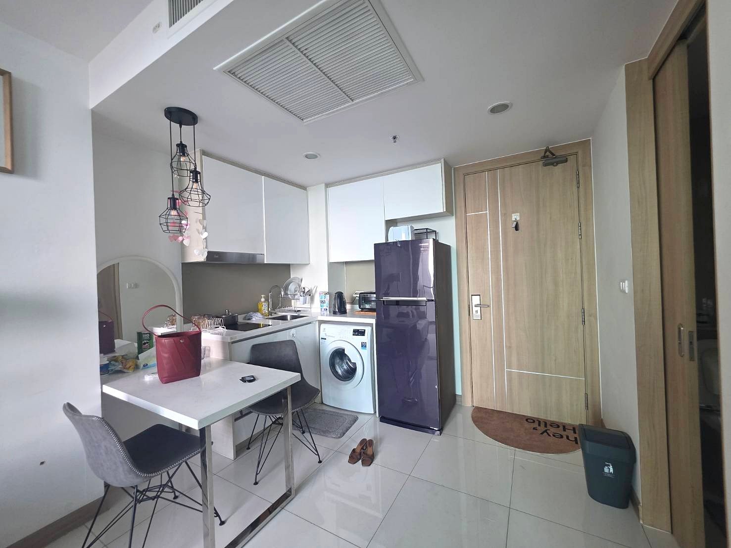 Riviera_Wongamat_B1209_7 Compact open-plan kitchen with white cabinets, a purple fridge, washing machine, and a small dining table with two chairs, in a bright space with hanging pendant lights.