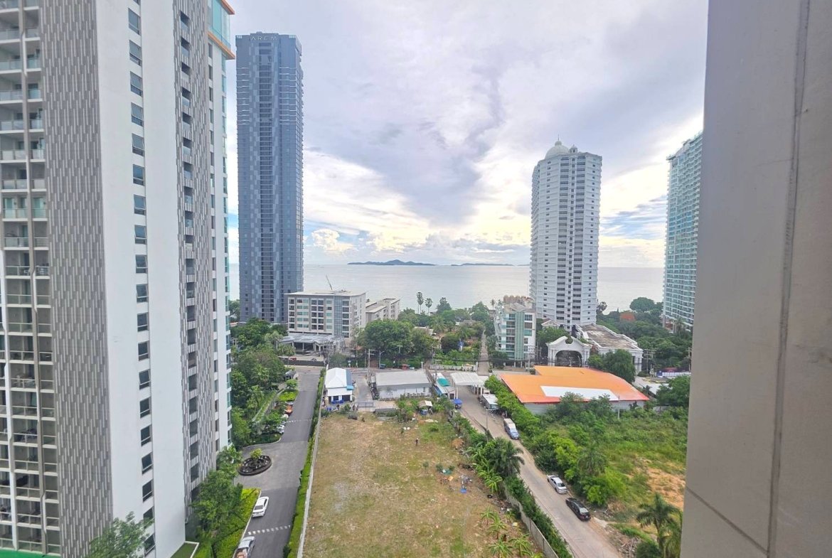City skyline with tall modern high-rise buildings along the coast, the sea and distant islands in the background, and a grassy lot with roads below.