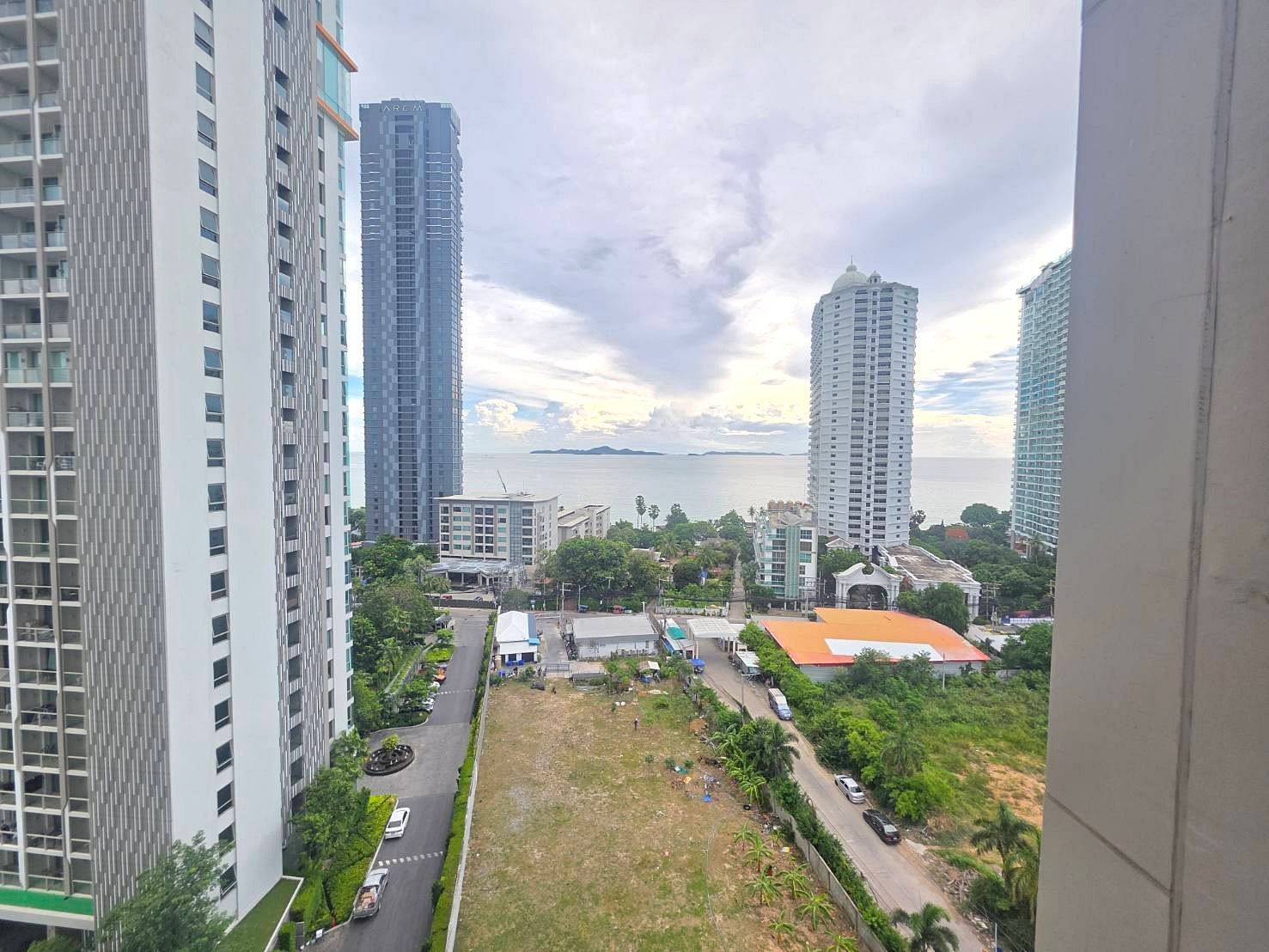 Riviera_Wongamat_B1209_9 City skyline with tall modern high-rise buildings along the coast, the sea and distant islands in the background, and a grassy lot with roads below.