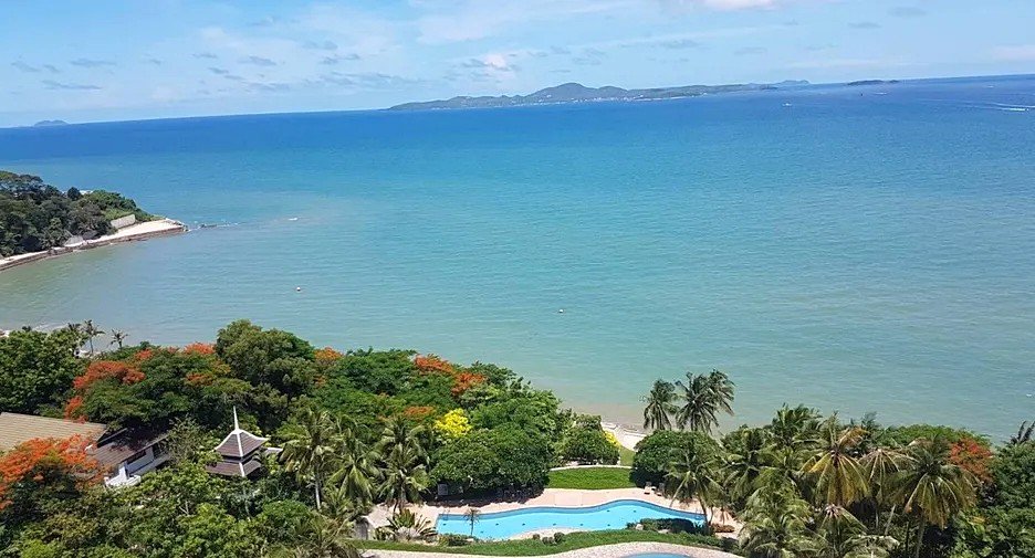 Aerial view of turquoise sea meeting a sandy beach, with a curving blue pool surrounded by palm trees and tropical greenery.