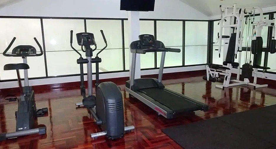 Interior of a bright gym with cardio machines—two treadmills, stationary bikes, and an elliptical—along polished wooden floors and large windows in the background.