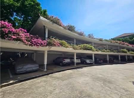 Two-level open parking deck with cars parked beneath a curved roof, and pink flowering plants along the railing against a blue sky.