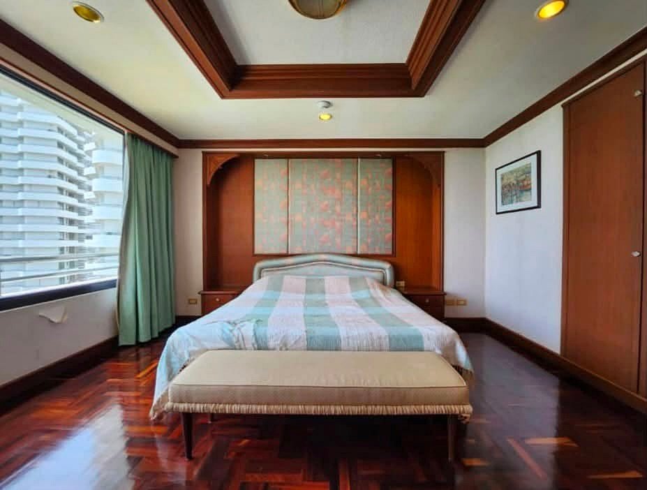 Hotel-style bedroom with a large bed, pastel quilt, beige bench at the foot, and wood-paneled headboard with built-in wardrobes and a window with green curtains.