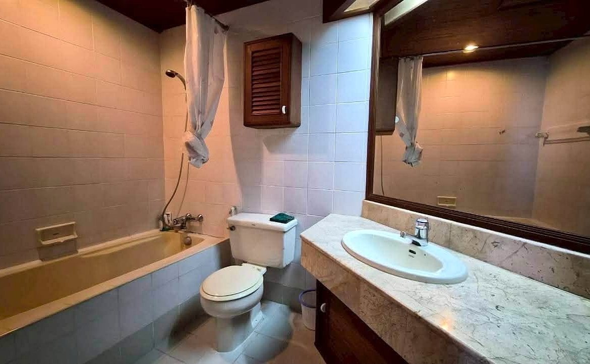 Bathroom with a tub/shower combo, toilet, and marble countertop sink beneath a large mirror with a tied curtain.