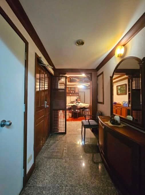 Narrow hallway in a traditional home with wooden doors and paneling, leading to a dining room at the far end. An arched mirror and sideboard are on the right, with warm lighting.