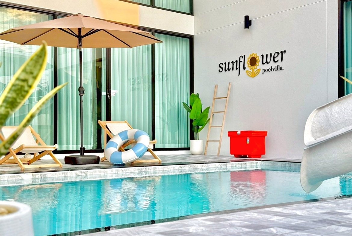 Poolside area at Sunflower Pool Villa: two wooden lounge chairs, a beige umbrella, lifebuoy, and a red cooler against a light wall.