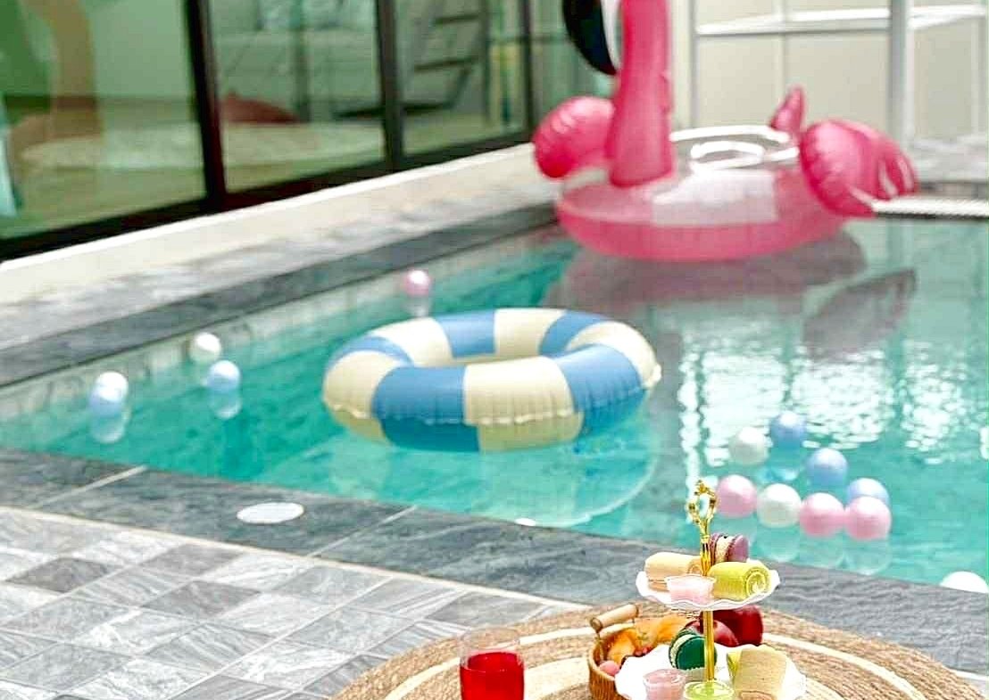 Poolside scene with a pink flamingo inflatable, a blue-and-yellow life ring in the pool, and a tiered dessert stand on a woven rug next to the water.