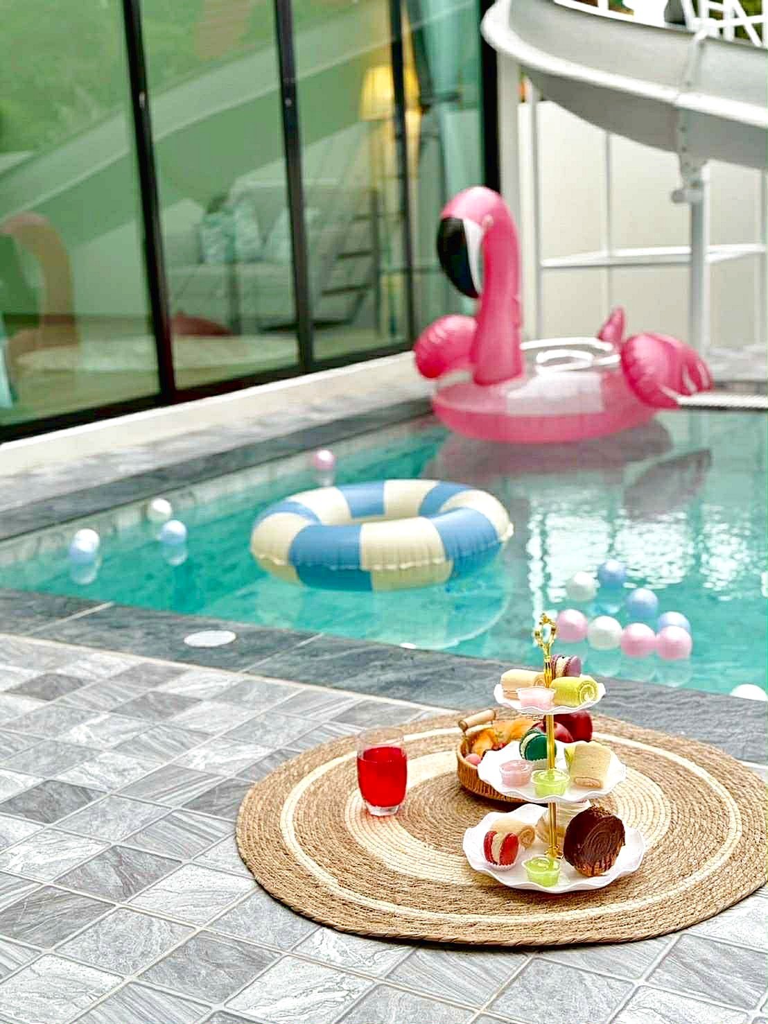 Sunflower_Pool_Villa_10 Poolside scene with a pink flamingo inflatable, a blue-and-yellow life ring in the pool, and a tiered dessert stand on a woven rug next to the water.