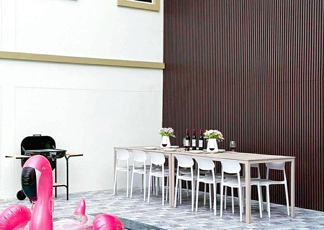 Poolside scene with a pink flamingo float in the pool, a long dining table set with bottles and flowers, white chairs, and a dark wood-paneled wall.