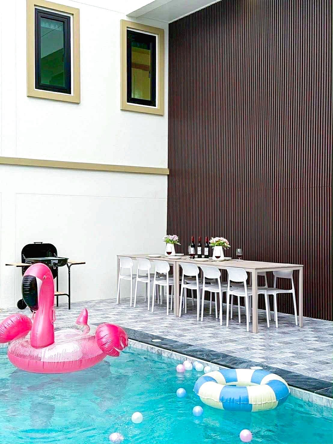 Sunflower_Pool_Villa_11 Poolside scene with a pink flamingo float in the pool, a long dining table set with bottles and flowers, white chairs, and a dark wood-paneled wall.