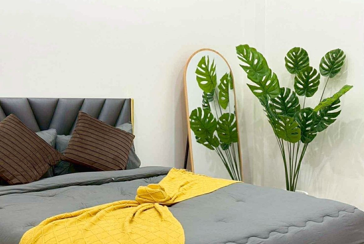 Bedroom scene with a gray upholstered bed, brown striped pillows, and a bright yellow quilt at the foot, plus a leaning mirror and large monstera plants nearby.