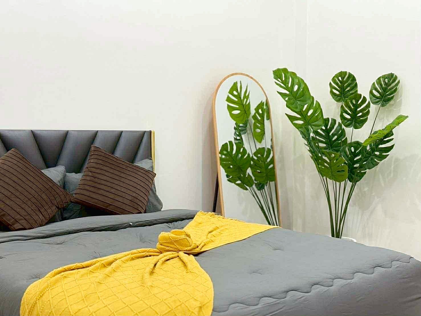 Sunflower_Pool_Villa_13 Bedroom scene with a gray upholstered bed, brown striped pillows, and a bright yellow quilt at the foot, plus a leaning mirror and large monstera plants nearby.