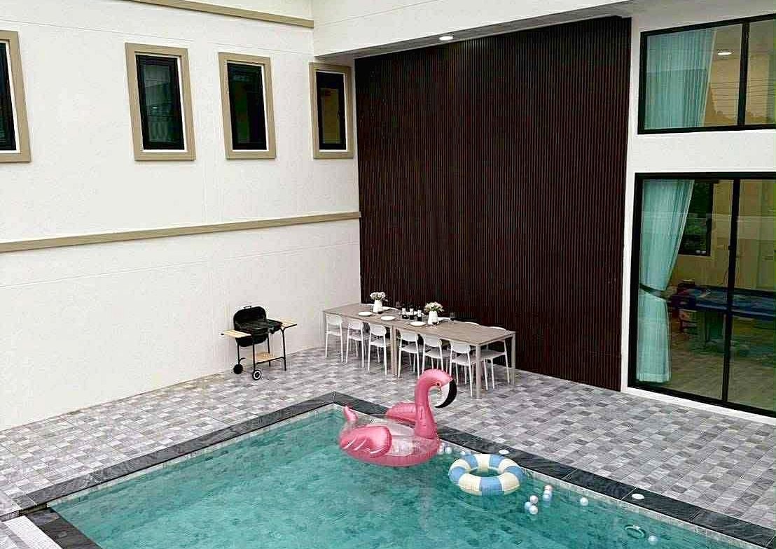 Modern poolside courtyard with a rectangular pool, pink flamingo float, and a white dining table set against a dark wood wall.