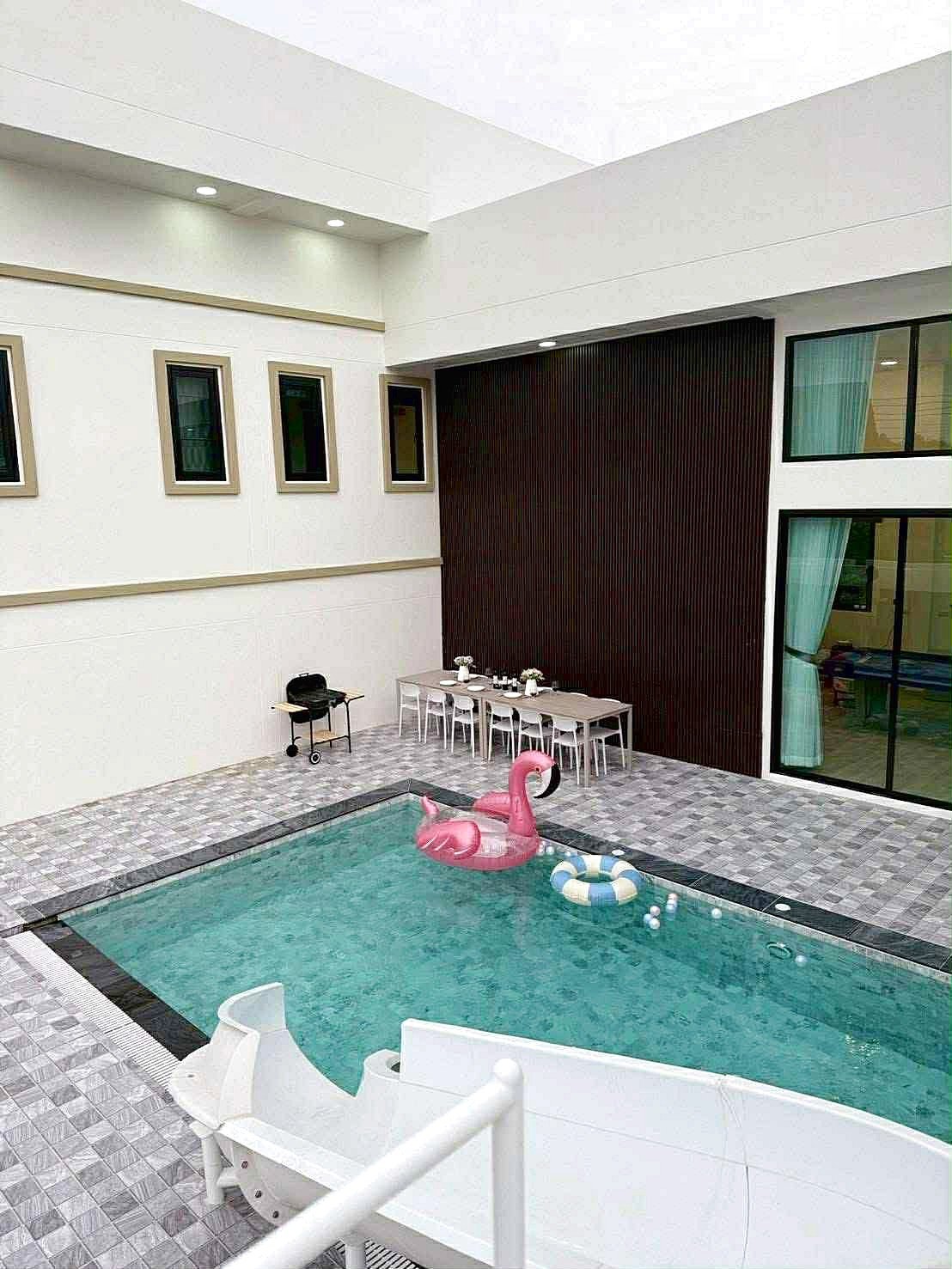 Sunflower_Pool_Villa_15 Modern poolside courtyard with a rectangular pool, pink flamingo float, and a white dining table set against a dark wood wall.