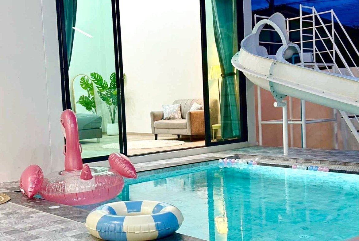 Outdoor pool scene with pink flamingo inflatable float and a blue-and-cream inner-ring float, near a glass-walled house with living room visible inside.