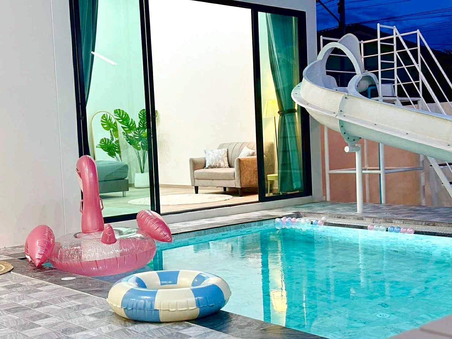 Sunflower_Pool_Villa_16 Outdoor pool scene with pink flamingo inflatable float and a blue-and-cream inner-ring float, near a glass-walled house with living room visible inside.