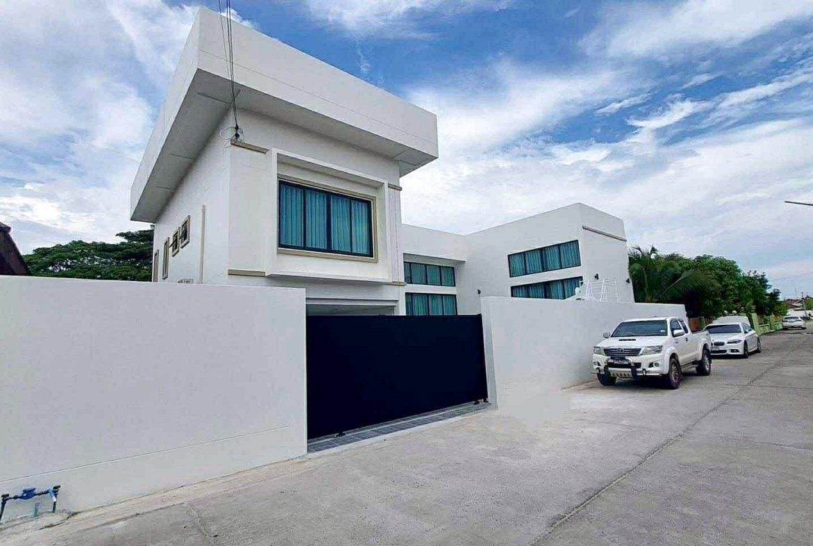 White modern house with a tall geometric facade and large windows, on a quiet street with parked white SUVs and a white wall gate in front