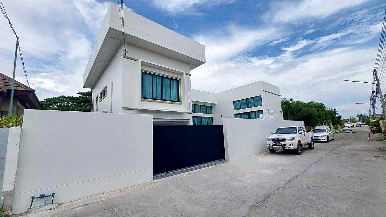 Sunflower_Pool_Villa_19 White modern house with a tall geometric facade and large windows, on a quiet street with parked white SUVs and a white wall gate in front