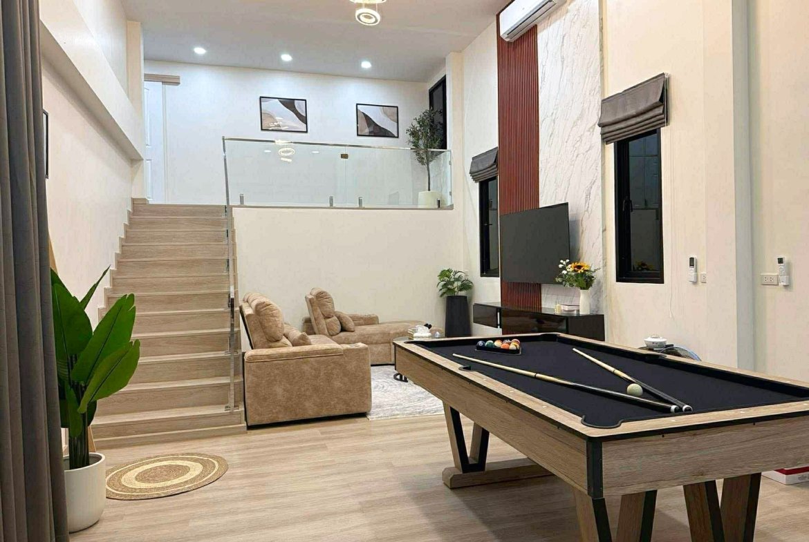 Modern living room with a pool table in the foreground, beige seating, staircase on the left, glass railing, and wall art above the stairs.