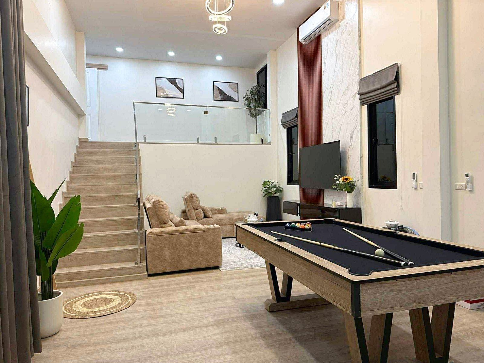 Sunflower_Pool_Villa_2 Modern living room with a pool table in the foreground, beige seating, staircase on the left, glass railing, and wall art above the stairs.