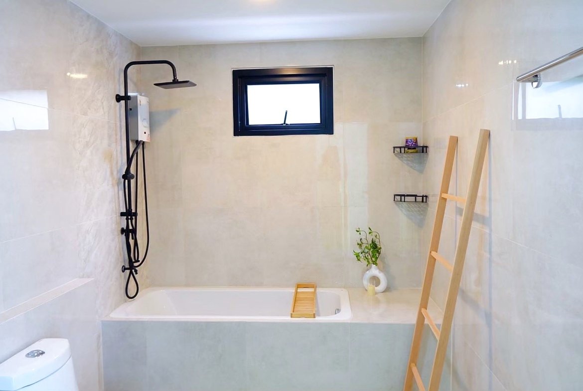 Bright minimalist bathroom with a white built-in tub, black ceiling-mounted rain shower, and a small rectangular window above the tub; a wooden bath tray rests on the tub and a wooden ladder leans against the wall near two corner shelves and a small plant in a white vase are visible on the tiled walls.