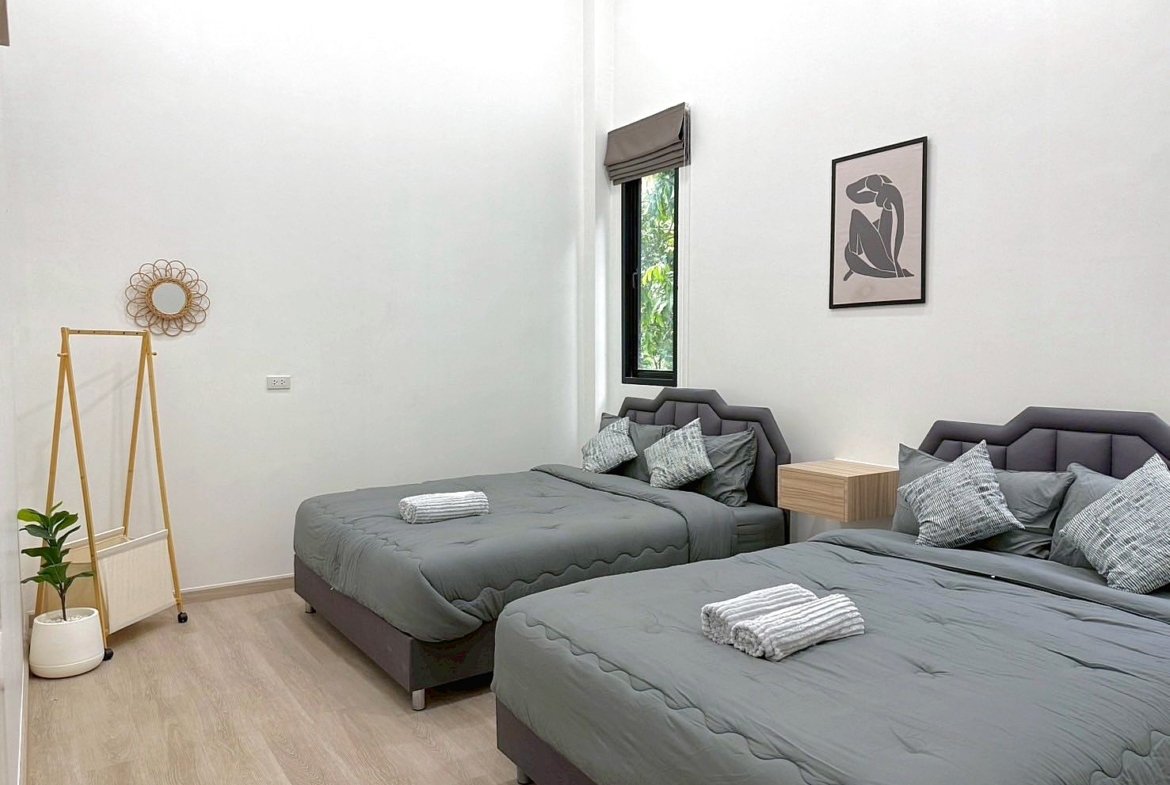 Twin bedroom with two gray-quilted beds, matching headboards, a wooden nightstand, and a window with a blind; minimalist decor and a potted plant nearby