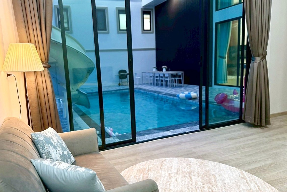 Living room with a beige sofa and blue cushions, a floor lamp, and large glass doors opening to a pool courtyard outside.
