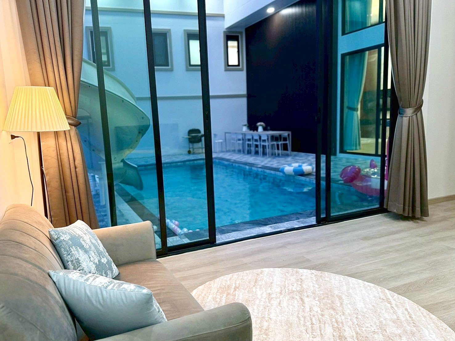 Sunflower_Pool_Villa_8 Living room with a beige sofa and blue cushions, a floor lamp, and large glass doors opening to a pool courtyard outside.