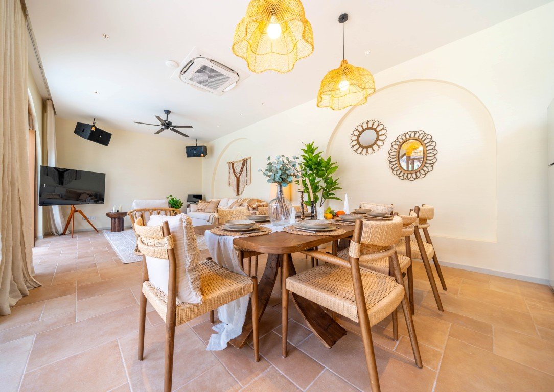 Bright open-plan dining room with a wooden table set for eight, woven chairs, and greenery under warm pendant lights