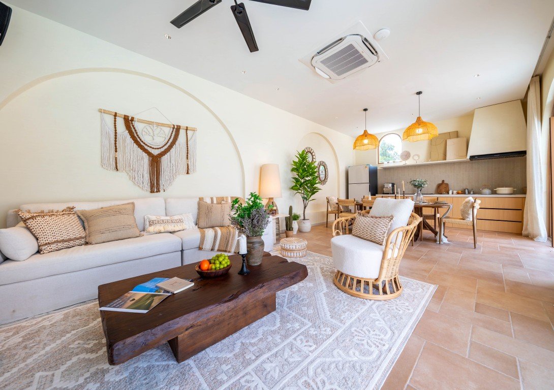 Bright open-plan living room with beige sofa, woven wall art, and a dark wood coffee table, extending into a kitchen area with pendant lights.