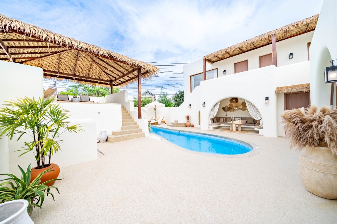 White Mediterranean-style villa with a curved blue pool, thatched shade structures, and a lounge area.
