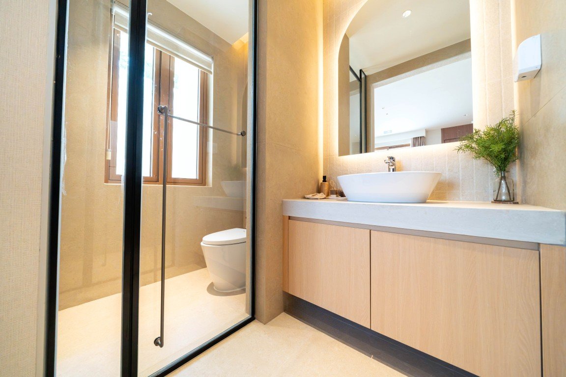 Modern bathroom with a glass shower enclosure, toilet, and a wooden vanity with a white vessel sink. A small plant sits by the sink under a large mirror.
