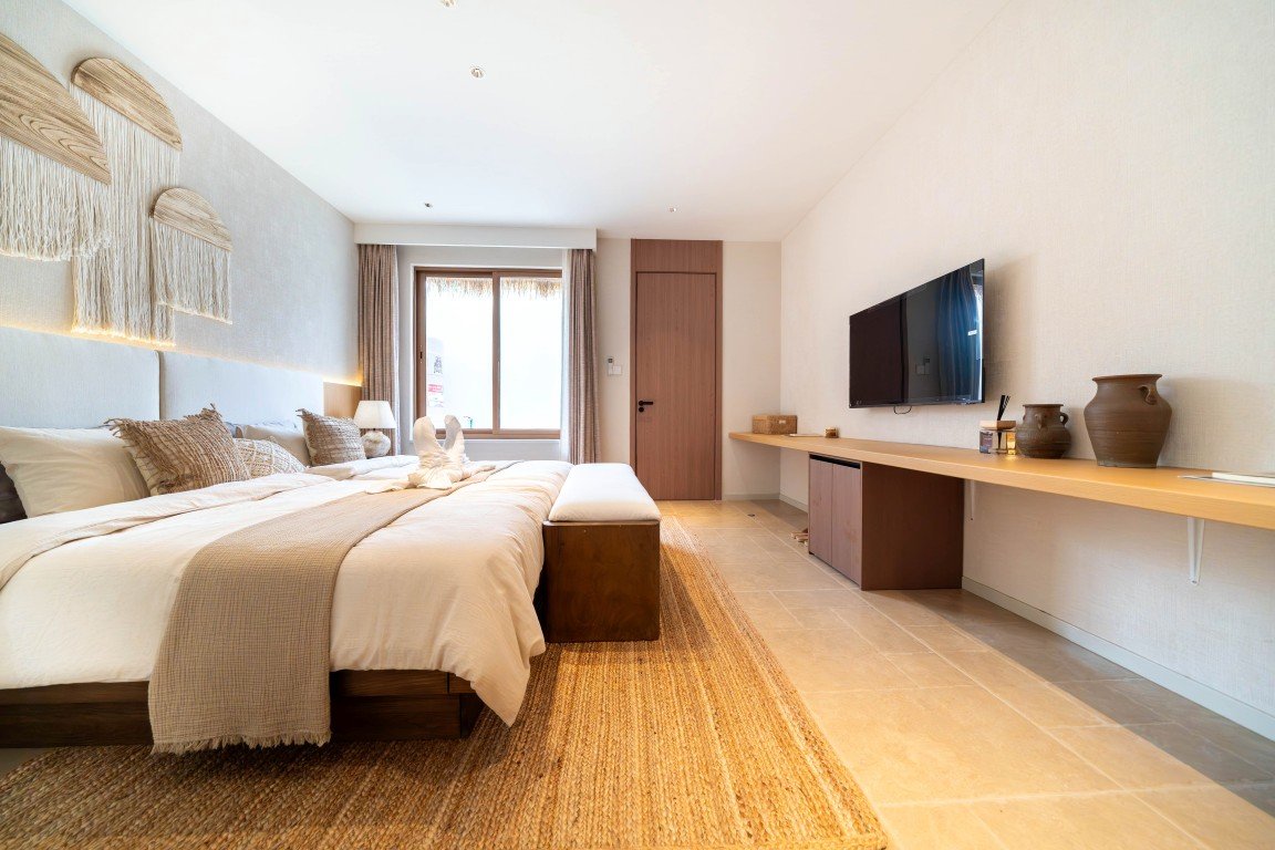 Hotel bedroom with a large bed, beige linens, and a woven rug; a bench at the foot and a wall-mounted TV opposite a long desk.
