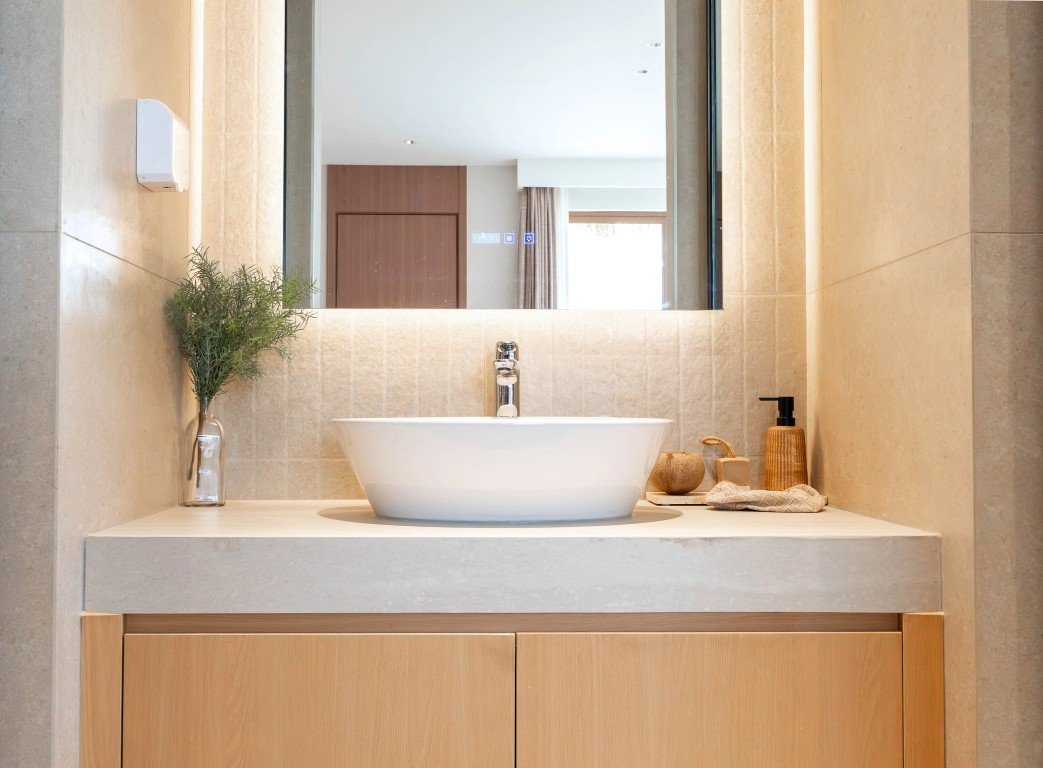 White round vessel sink on a beige stone countertop with a tall mirror, wooden cabinets, and a potted plant on the left.
