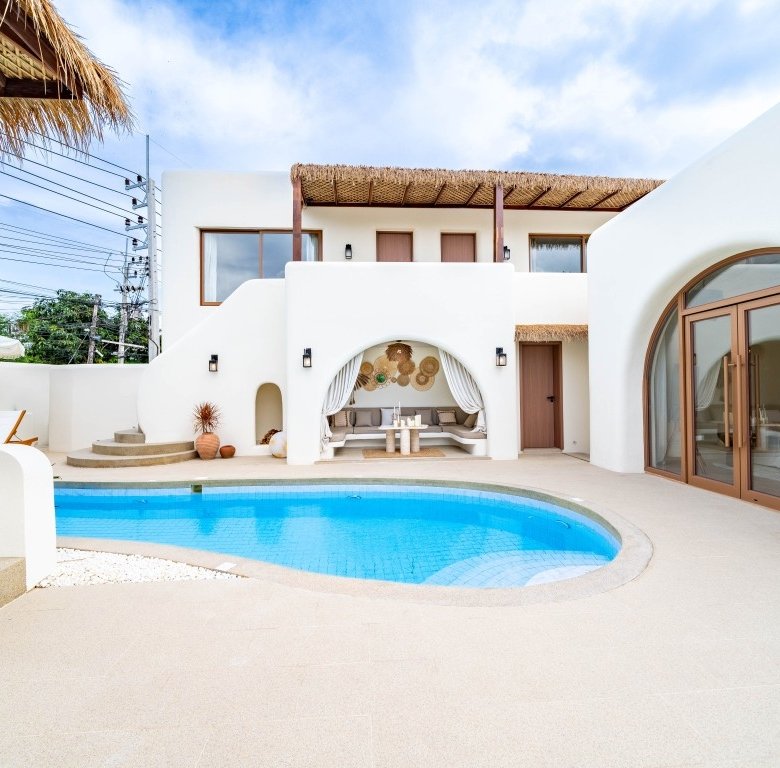 White washed villa courtyard with a curved blue pool and a shaded seating alcove under a thatched canopy