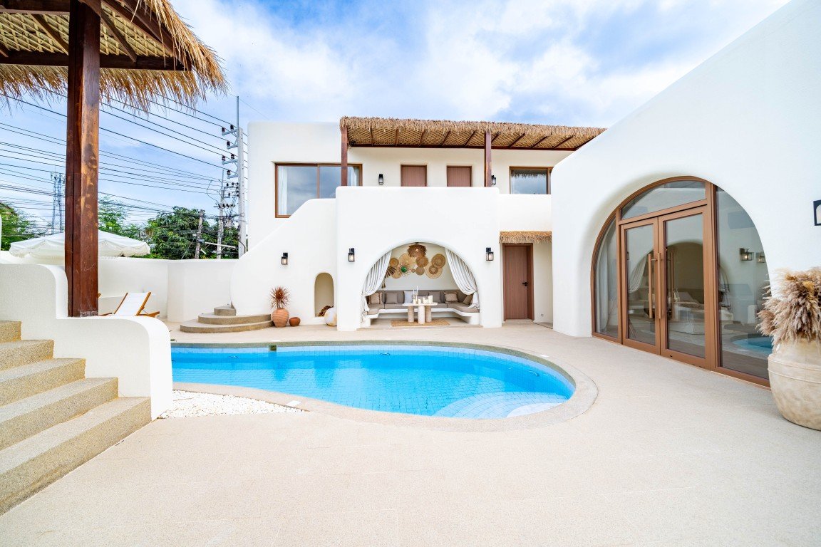 White washed villa courtyard with a curved blue pool and a shaded seating alcove under a thatched canopy