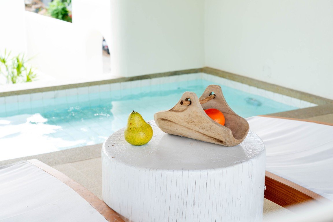 Pear resting on a white cylindrical table next to a wooden fruit holder with an orange, by a bright indoor pool area