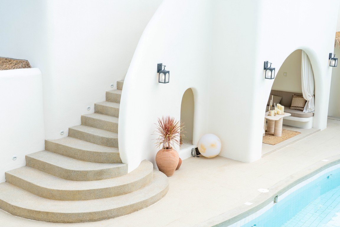 Curved white Mediterranean-style wall with stairs beside a pool and a terracotta vase with dried plant by the doorway.