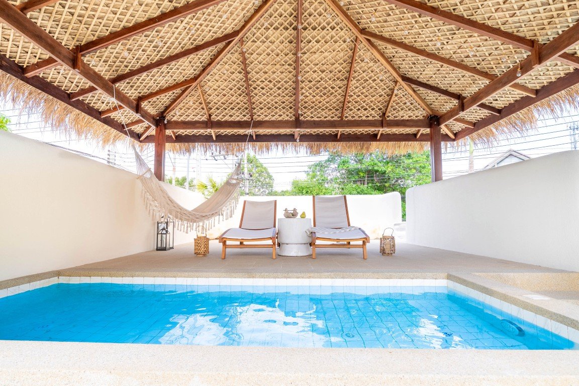 Poolside patio with a thatched roof structure, two white lounge chairs, a hammock, and lanterns overlooking a blue swimming pool in the foreground