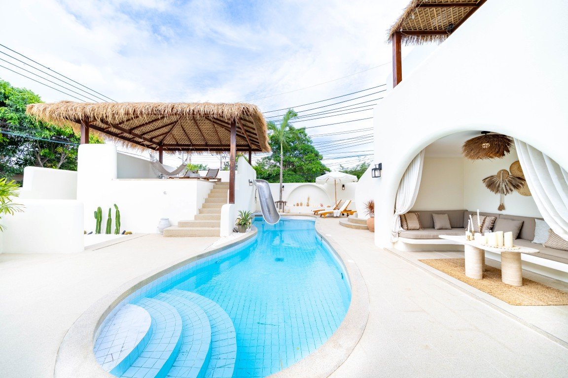 Curved turquoise pool in a sunlit white villa with thatched cabanas and a shaded lounge area nearby.