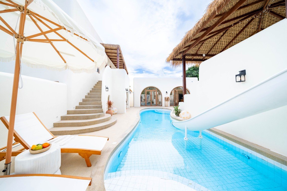 Curved blue pool bordered by white walls and thatched shade structures, with sun loungers and an umbrella nearby.
