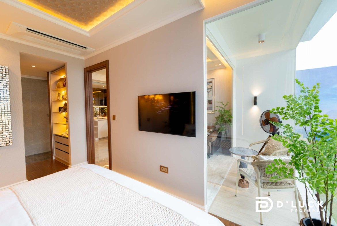 Hotel bedroom with a wall-mounted flat-screen TV, beige walls, and a glass door leading to a balcony with seating and potted plants. The bed is in the foreground with white linens.