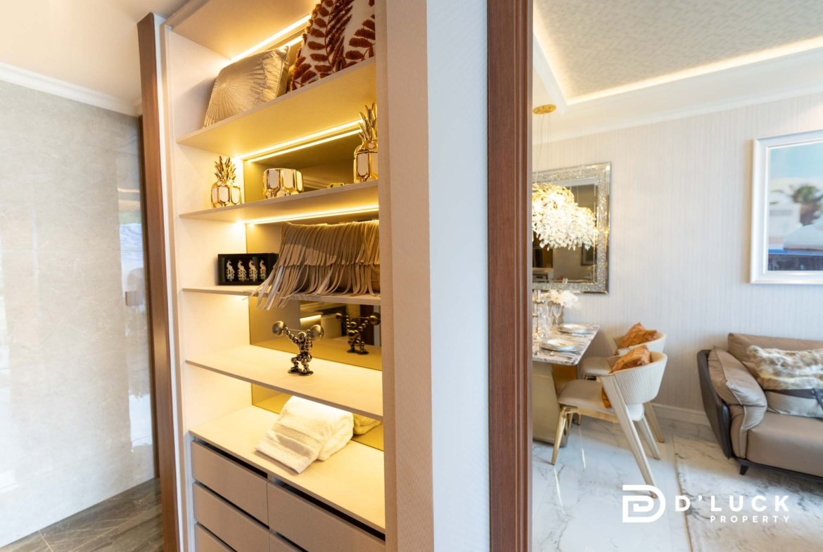 Open built-in wardrobe with illuminated shelves displaying gold pineapple decor, towels, and hanging items; dining area visible beyond.